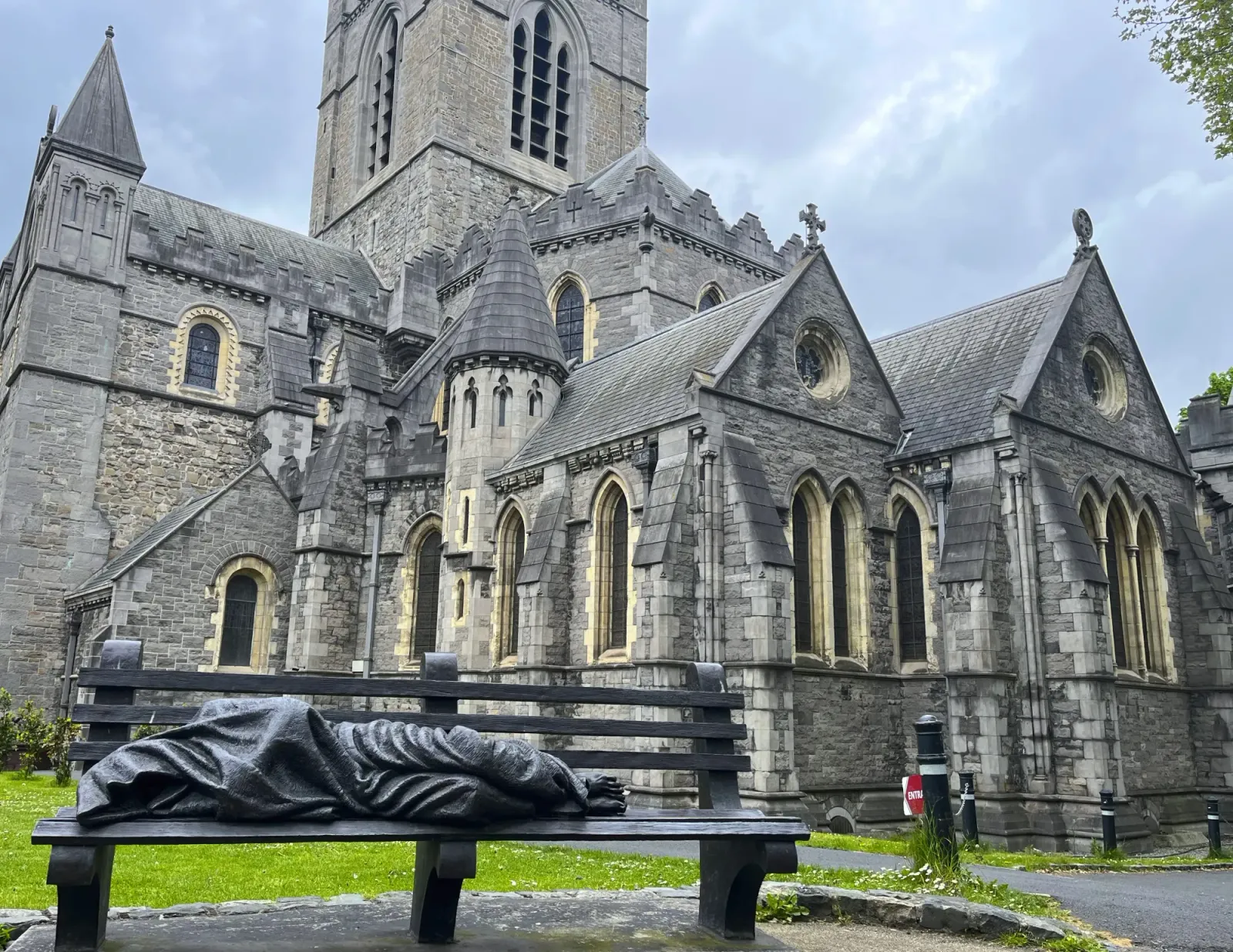 Christ Church Cathedral by Mr. Wanderer Statue of a wrapped figure laying on a bench in front of a grey stone cathedral