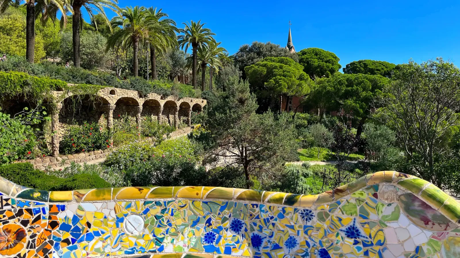 Park Güell by Mrs. Wanderer Tiled balcony overlooking a park with arches and palm trees