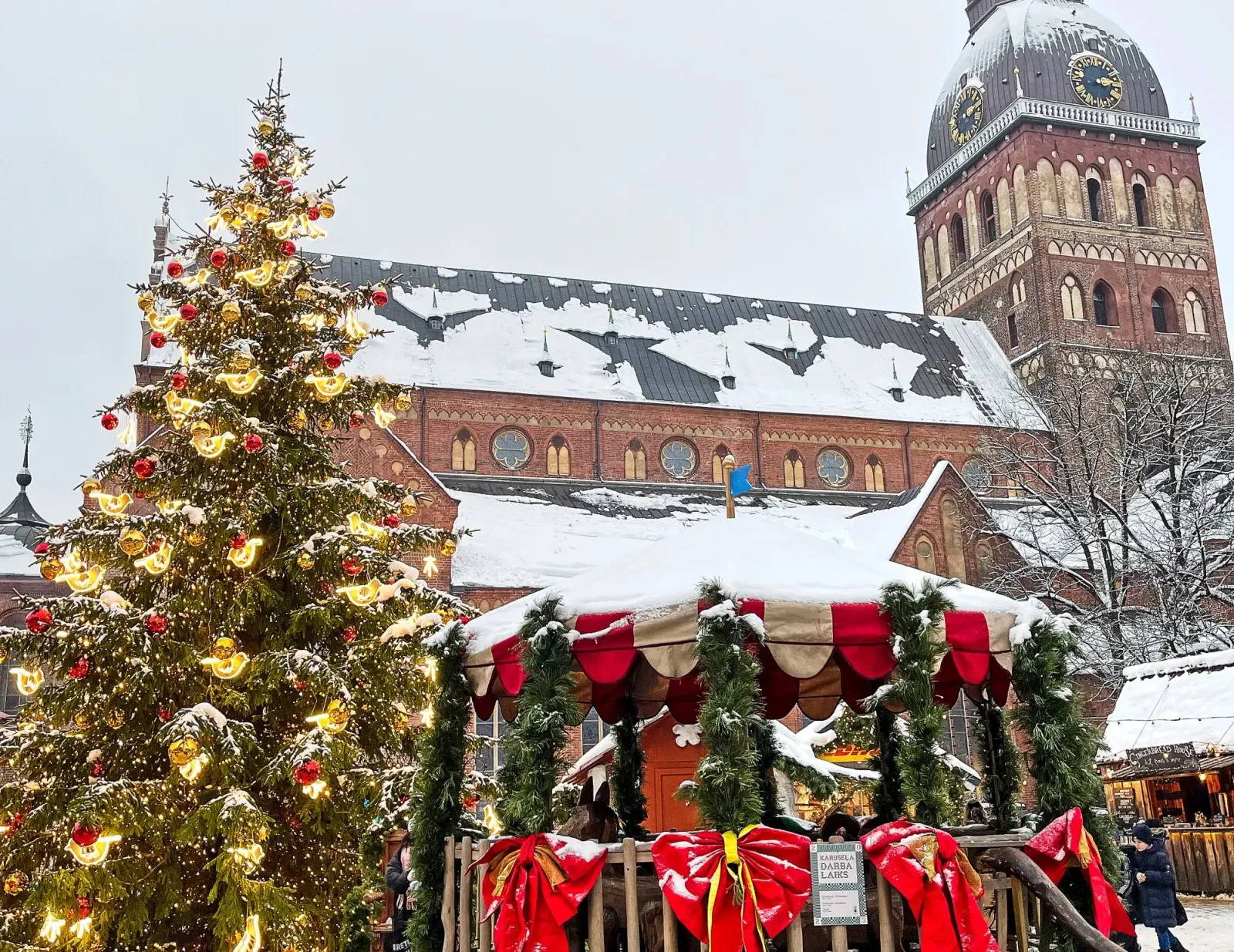 Riga Christmas Market by Mrs. Wanderer Tinsel tree and bedecked gazeebo in front of a brick church covered in snow