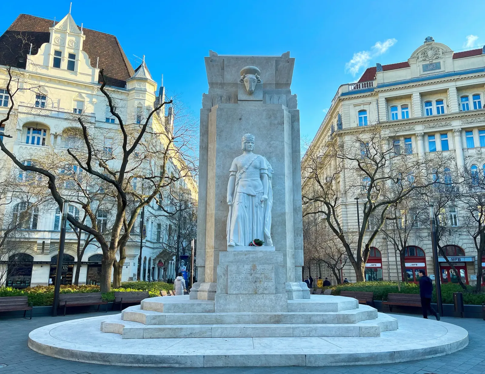 Monument of National Martyrs by Mr. Wanderer Stone rectangular column with a warrior on a pedastal resting in front of two cream-colored buildings