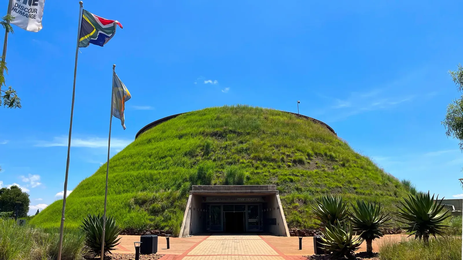 Cradle of Humankind by Mr. Wanderer Grass covered conical building with a gaping entrance and adjoining flags