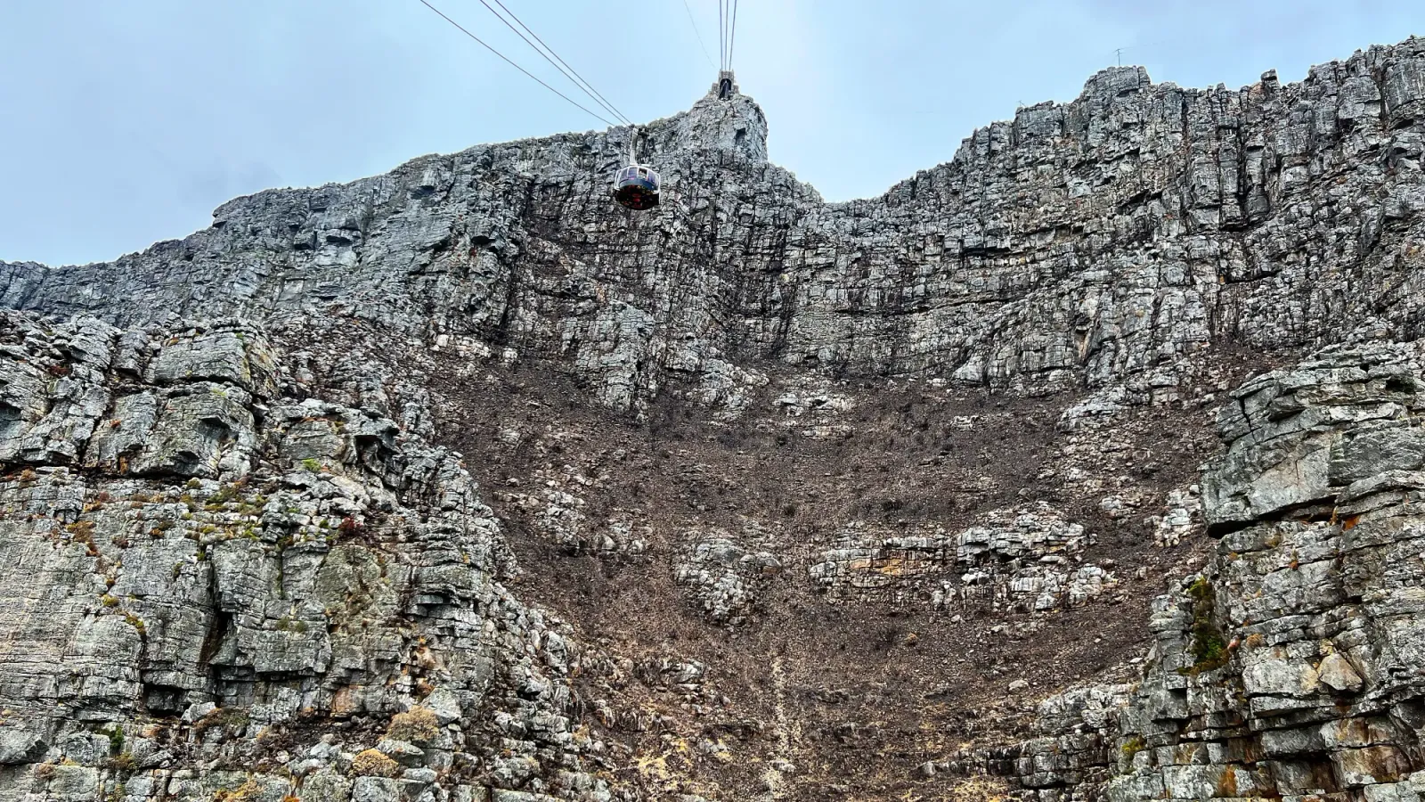 Table Mountain Cable Car by Mrs. Wanderer Rock face with a round cable car and accompanying wires draping from it