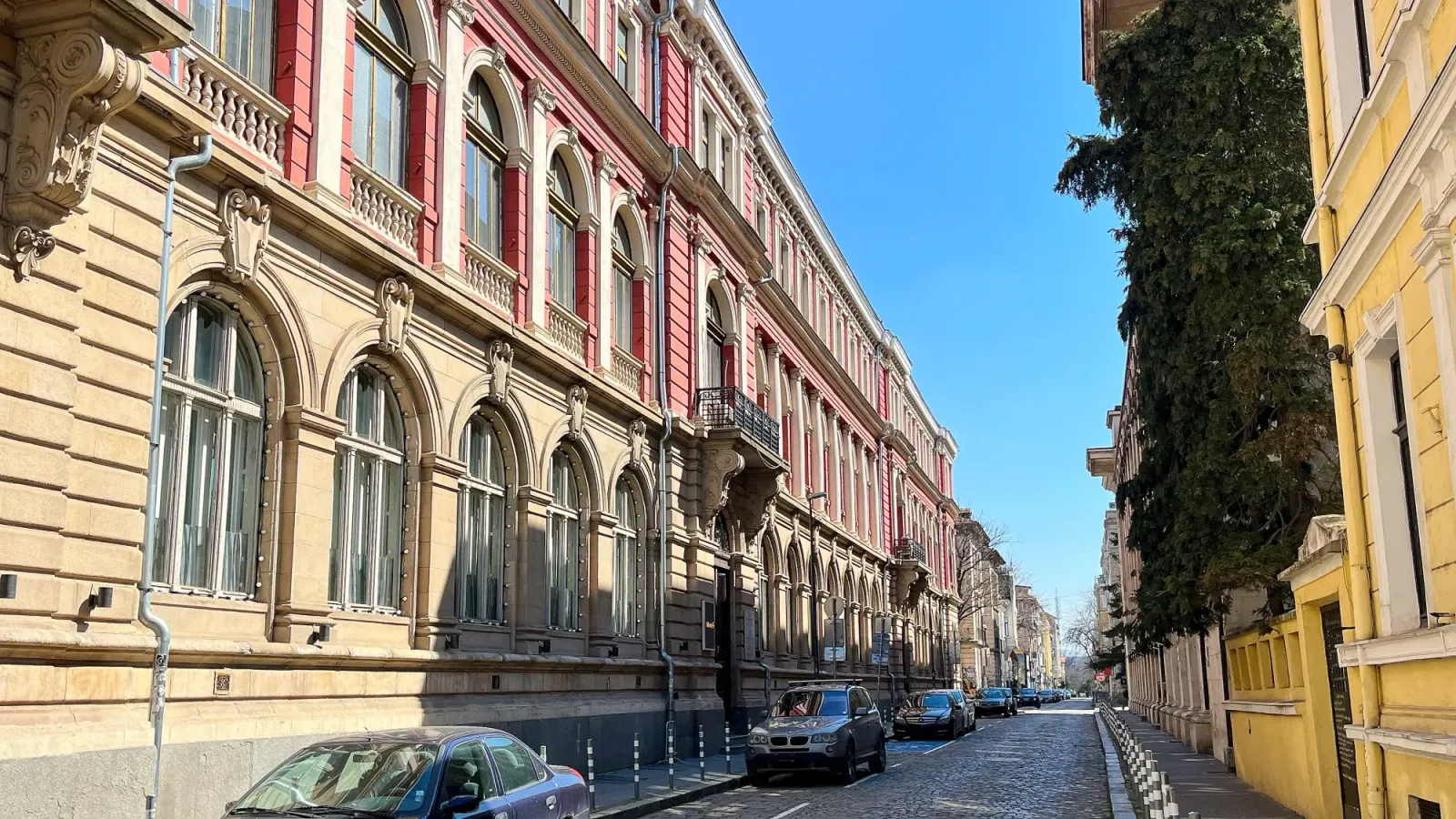 Sofia Streets by Mrs. Wanderer Stone streets with pastel buildings and a row of parked cars.