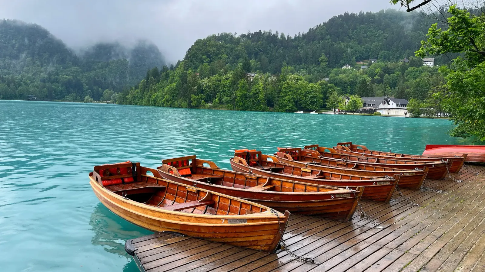Wooden Bled Boats by Mrs. Wanderer Six wooden boats docked against turquise water with greenery all around
