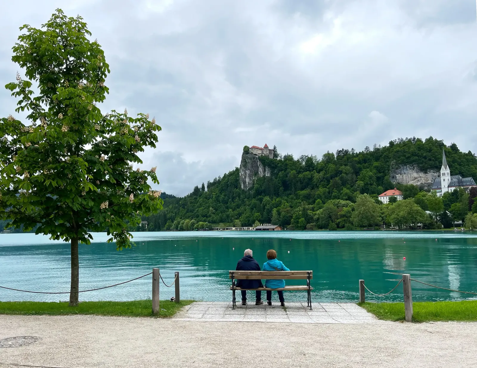 Watching Bled Castle by Mrs. Wanderer Two folks sitting on a bench looking out over the lake and the castle on the opposite shore