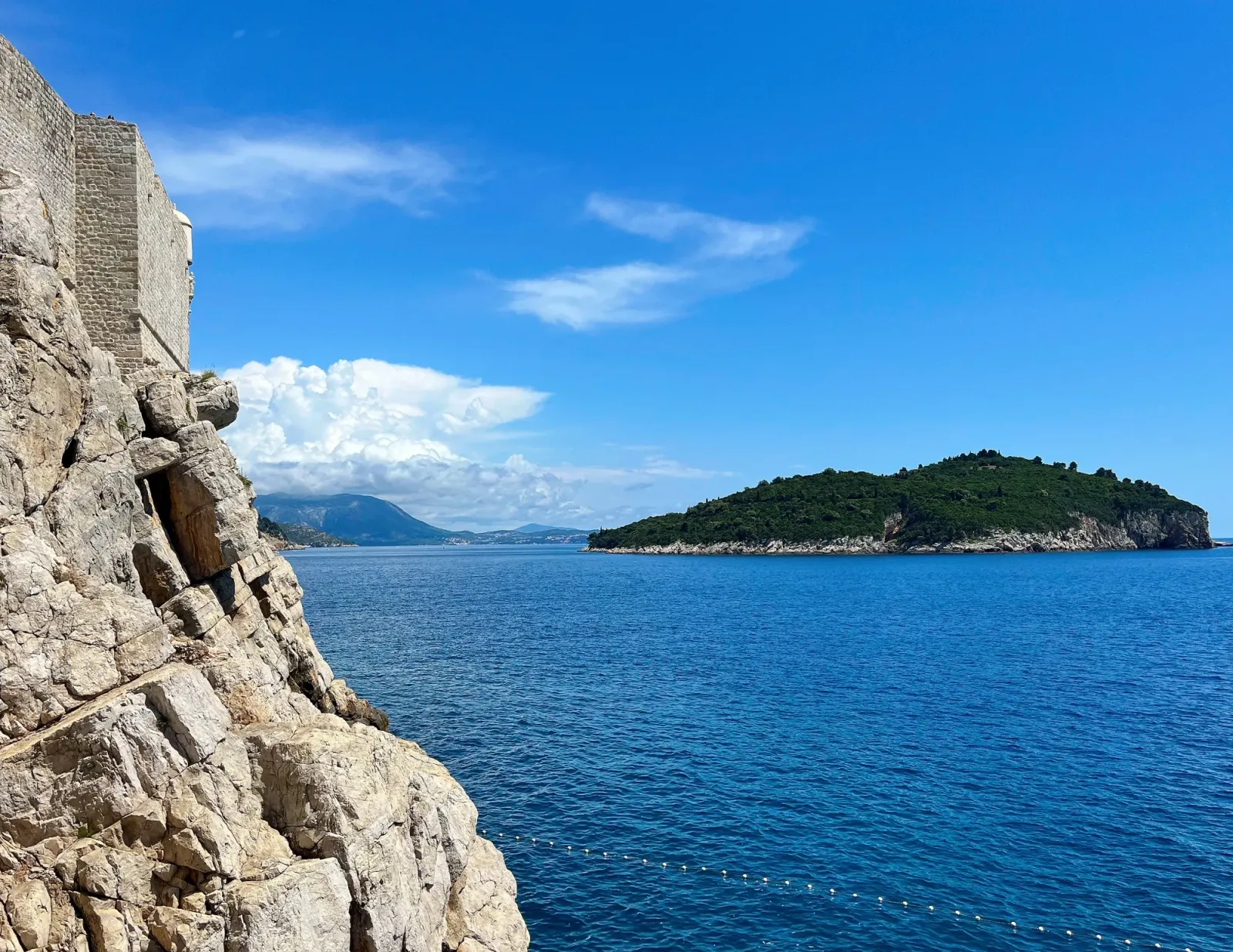 Dubrovnik City Walls by Mrs. Wanderer Stone walls set atop a stone cliff overlooking the blue sea with an island in the background