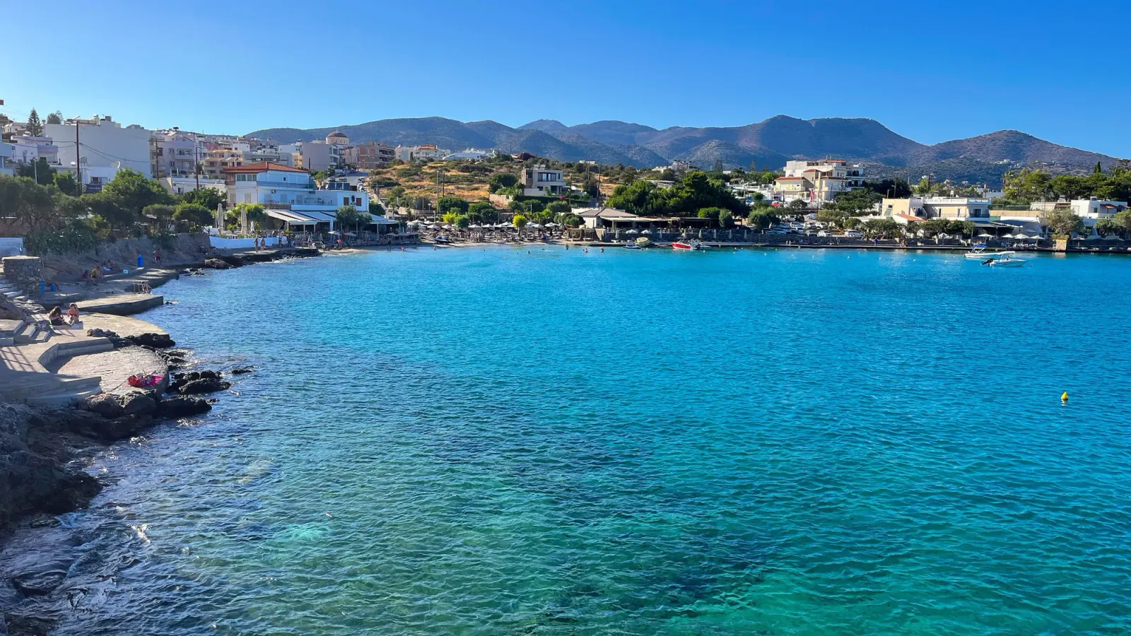 Cretan Seaside by Mrs. Wanderer Multi-shades of blue water with stone and sandy spots filled with sunbathers and swimmers