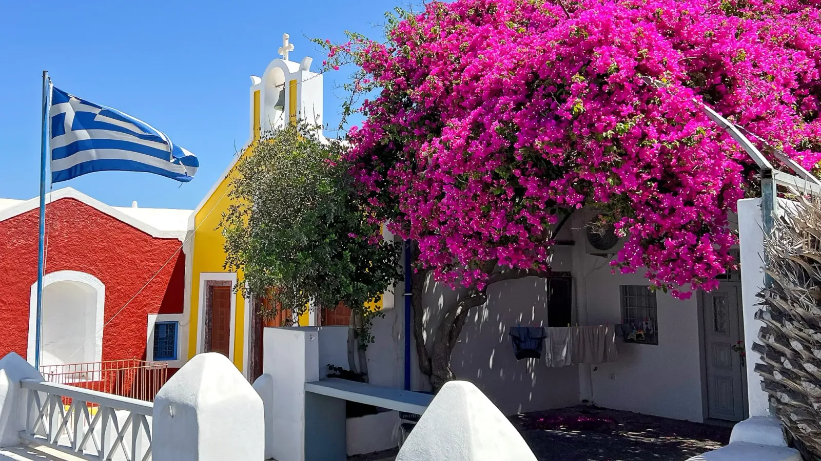 Greek Bougainvillea by Mr. Wanderer Flying greek flag, yellow church facade, with vibrant pink bougainvillea growing before them